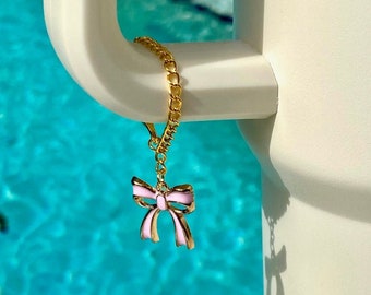 May include: A gold chain with a pink bow charm hanging from a white surface. The chain is hanging over a blue pool.
