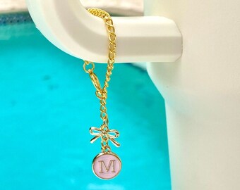May include: Gold-colored chain charm with a pink circular pendant featuring the letter "M" and a small bow. The charm is attached to a white handle, with a turquoise blue background.