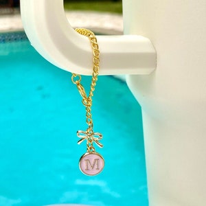 May include: Gold chain charm with a pink circular pendant bearing the letter "M" and a small bow. The charm is attached to a white handle, with a turquoise blue background.
