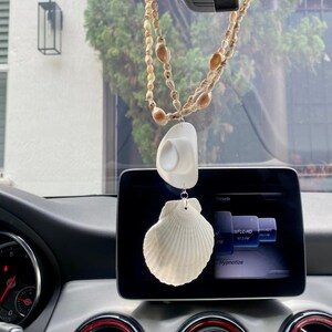 May include: A white seashell car charm hanging from a rearview mirror. The charm is attached to a string of small seashells. The car charm is hanging in a car with a black dashboard and a black rearview mirror.