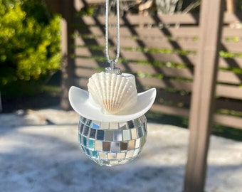 May include: A silver disco ball ornament with a white cowboy hat and a white seashell on top. The ornament is hanging from a silver chain.