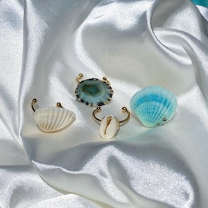 May include: Three gold rings with different seashell designs. The rings feature a white seashell, a blue seashell, and a green and white seashell.