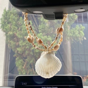 May include: A car air freshener made of a string of seashells with a large seashell at the bottom. The seashells are a light beige color. The air freshener is hanging from the rearview mirror of a car.
