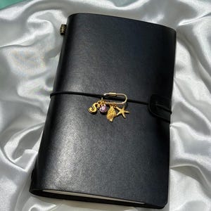 以下が含まれることがあります： 黒い革製のジャーナルで、黒いゴムバンドで留められています。バンドには、文字のS、小さなピンクの円形ジェム、質感のある金色の貝殻、そしてヒトデのチャームが付いた金色の安全ピン型のチャームが取り付けられています。