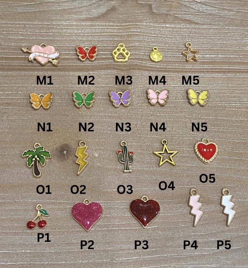 May include: A collection of gold-toned charms with colorful enamel details. Designs include butterflies, hearts, stars, a paw print, a palm tree, a cactus, cherries, and lightning bolts. Each charm is labeled with a letter and number combination.