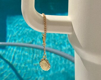 May include: A gold chain necklace with a seashell pendant. The necklace is hanging from a white ceramic mug. The mug is in front of a blue swimming pool.