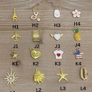 May include: A collection of gold-colored charms, including an Eiffel Tower, a stethoscope, a jar with a heart, a flower, an airplane, a $100 bill, a heart with wings, a pineapple, a star, a heart with "Mom" on it, a shirt with a heart, a star with a flag design, a sunburst, a seashell, a starfish, and a shell.