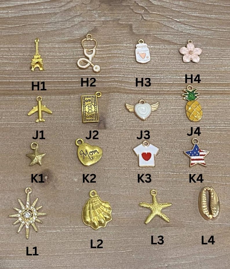 May include: A collection of gold-colored charms, including an Eiffel Tower, a stethoscope, a jar with a heart, a flower, an airplane, a $100 bill, a heart with wings, a pineapple, a star, a heart with "Mom" on it, a shirt with a heart, a star with a flag design, a sunburst, a seashell, a starfish, and a shell.