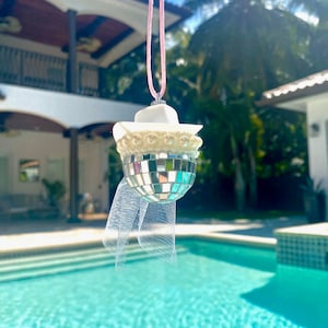 May include: A white disco ball ornament with a white hat and veil, hanging from a pink string. The ornament is in front of a swimming pool.