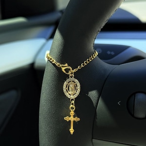 May include: A gold chain with a charm featuring a religious icon and a cross. The chain is hanging from the headrest of a car.