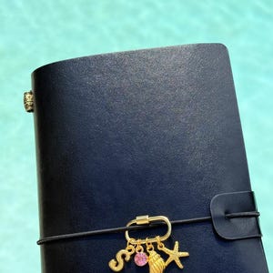 以下が含まれることがあります： 黒いレザー調のジャーナルで、黒いゴムバンドで留められています。バンドにはゴールドトーンのカラビナチャームが付いており、ゴールドの文字S、小さな丸いピンクのクリスタル、質感のあるゴールドの貝殻、ゴールドのヒトデのチャームがぶら下がっています。