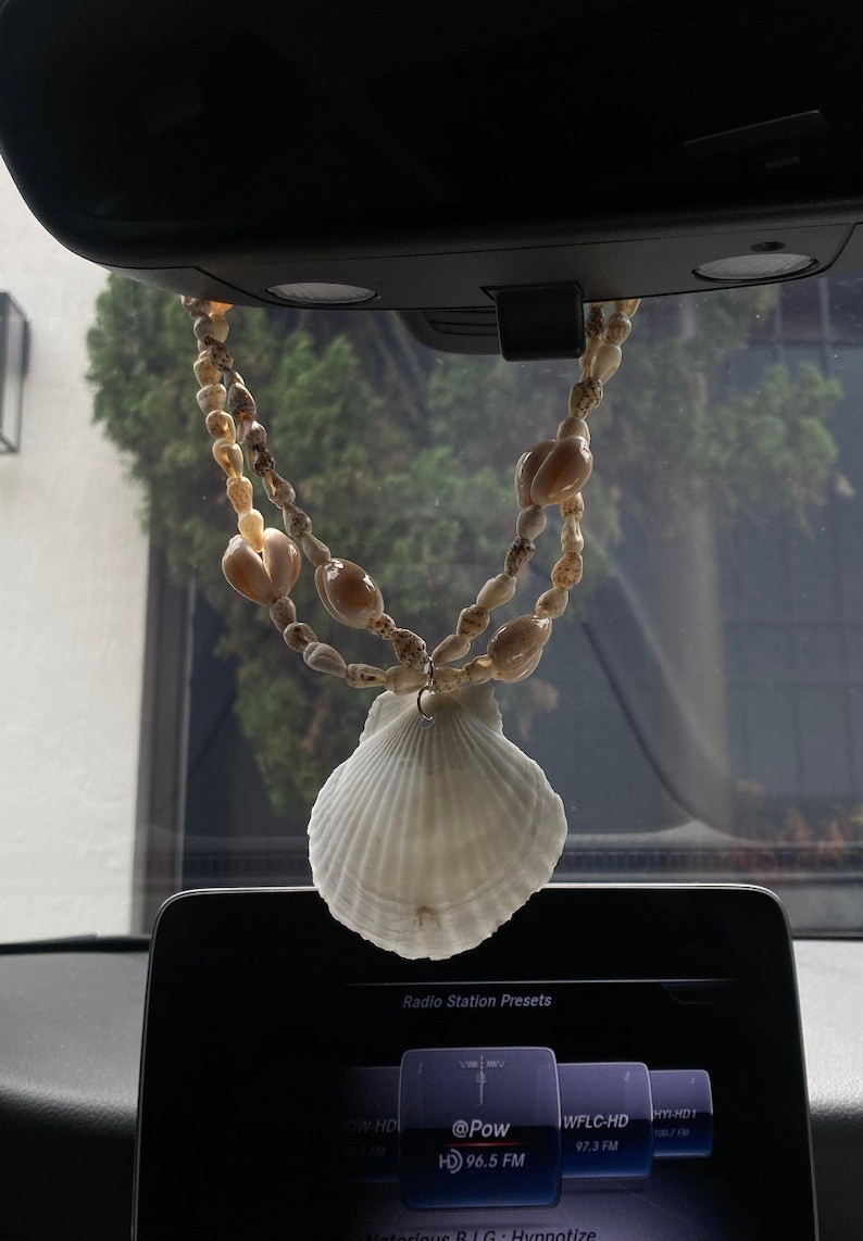 Puede incluir: Una concha blanca colgando del espejo retrovisor de un coche. La concha est&aacute; unida a una cuerda de peque&ntilde;as conchas marrones.