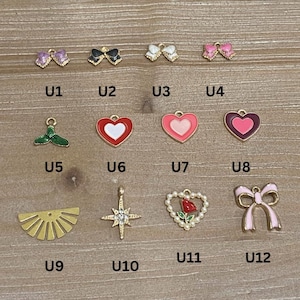 May include: A collection of small, colorful charms in various shapes, including bows, hearts, and a sunburst design. The charms are gold-toned and feature enamel details in pink, red, black, and green. Each charm has a small loop for attaching to jewelry.