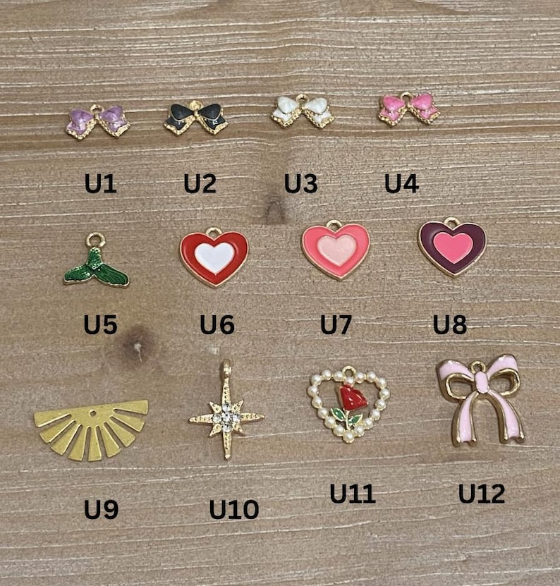 May include: A collection of small, colorful charms in various shapes, including bows, hearts, and a sunburst design. The charms are gold-toned and feature enamel details in pink, red, black, and green. Each charm has a small loop for attaching to jewelry.