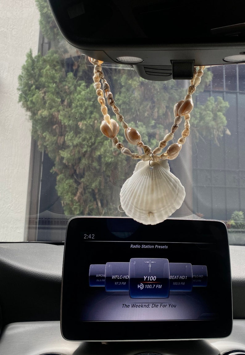 Puede incluir: Un espejo retrovisor de un coche con una guirnalda de conchas colgando de &eacute;l. La guirnalda est&aacute; hecha de conchas blancas y marrones. La pantalla de radio del coche muestra la estaci&oacute;n "Y100" reproduciendo "The Weeknd: Die For You".