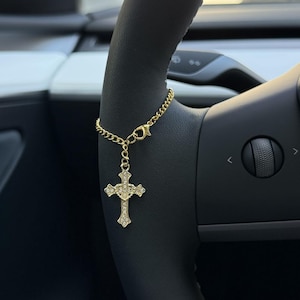 May include: A gold-colored cross charm with clear crystals hangs from a gold chain attached to a car steering wheel. The cross is ornate, with a detailed design. The steering wheel is black.
