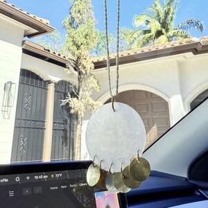 May include: A white, round, shell-shaped car charm with a string hanging from the top. The charm has a gold ring around the edge and gold, round, metal bells hanging from the bottom.