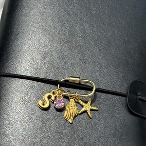 以下が含まれることがあります： ゴールドトーンの金属製カラビナクリップ。ゴールドの文字「S」、丸いピンクのファセットカットクリスタル、質感のあるゴールドの貝殻、ゴールドのヒトデという4つのチャームがぶら下がっています。クリップは細い黒のゴムバンドに取り付けられています。