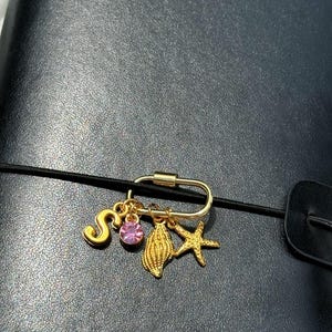 以下が含まれることがあります： 黒いゴムバンドに取り付けられたゴールドトーンの金属製カラビナクリップ。ゴールドの文字「S」、丸いピンクのラインストーン、質感のあるゴールドの貝殻、ゴールドのヒトデという4つのチャームがぶら下がっています。チャームは一列に並んでいます。