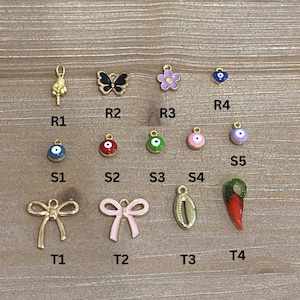 May include: A collection of gold-toned charms and pendants, including a butterfly, flower, bow, and various eye-shaped designs. Other charms include a hand gesture, a shell, and a red chili pepper. The charms are arranged on a light wood surface.