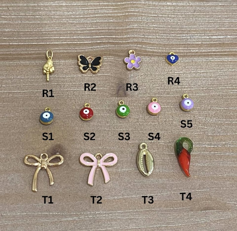 May include: A collection of gold-toned charms and pendants, including a butterfly, flower, bow, and various eye-shaped designs. Other charms include a hand gesture, a shell, and a red chili pepper. The charms are arranged on a light wood surface.