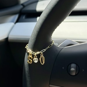 May include: A gold-colored charm bracelet with three dangling charms. The charms include the letter "S", a clear crystal, and the number "0". The bracelet is hanging on a black steering wheel.