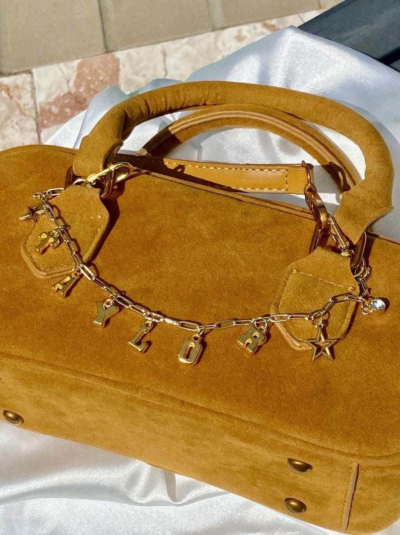 May include: A tan suede handbag with a rounded top handle and gold-colored hardware. A gold chain with the letters "Y L O R" and a star charm hangs from the bag. The bag has a structured shape and a soft texture.