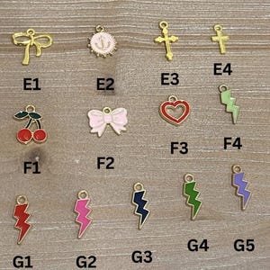 以下が含まれることがあります： E1からG5のラベルが付いた小さな金属製チャームのコレクション。金色のリボン、ピンクのアンカー付き円形、金色の十字架、赤いサクランボのペア、ピンクのリボン、赤いハート、そして緑、紫、赤、ピンク、紺色の稲妻の形をしたチャームが含まれています。