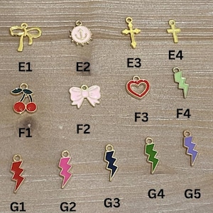 May include: A collection of gold-colored charms with various designs, including a bow, anchor, cross, cherries, a pink bow, a red heart, and lightning bolts in red, pink, navy, green, and purple. The charms are arranged on a wooden surface.