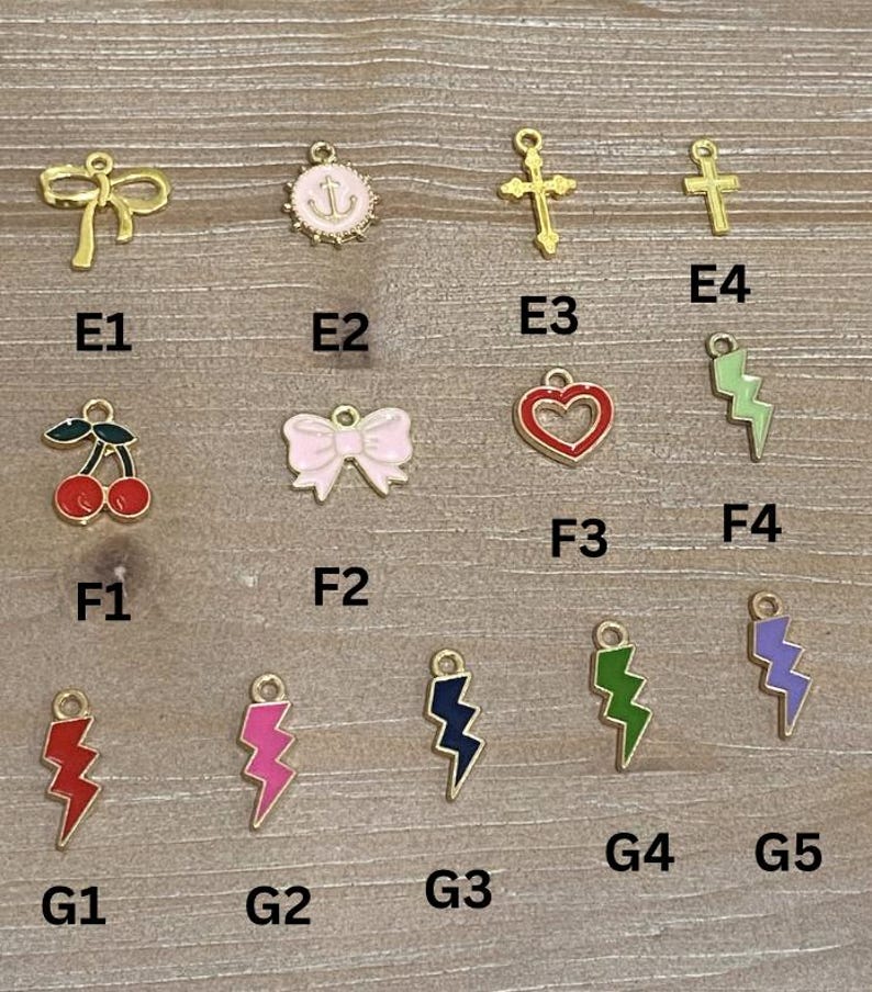 May include: A collection of gold-colored charms with various designs, including a bow, anchor, cross, cherries, a pink bow, a red heart, and lightning bolts in red, pink, navy, green, and purple. The charms are arranged on a wooden surface.