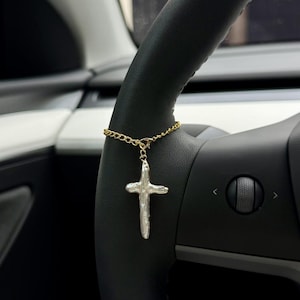 May include: A gold-colored chain with a white cross pendant hanging from a black steering wheel. The cross has a textured, irregular surface. The steering wheel is black with silver accents.
