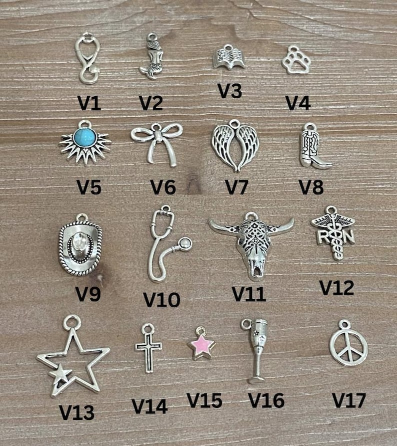May include: A collection of silver-toned charms, including a stethoscope, mermaid, angel wings, paw print, sunburst with turquoise center, bow, cowboy boot, star, cross, and peace sign. The charms are displayed on a wooden surface.