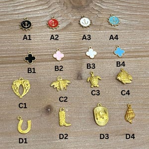 May include: A collection of gold-colored charms with various designs, including an anchor, clover, wings, bee, turtle, fish, horseshoe, cowboy boot, cowboy hat, and seashell. The charms have black, red, white, and blue enamel accents.