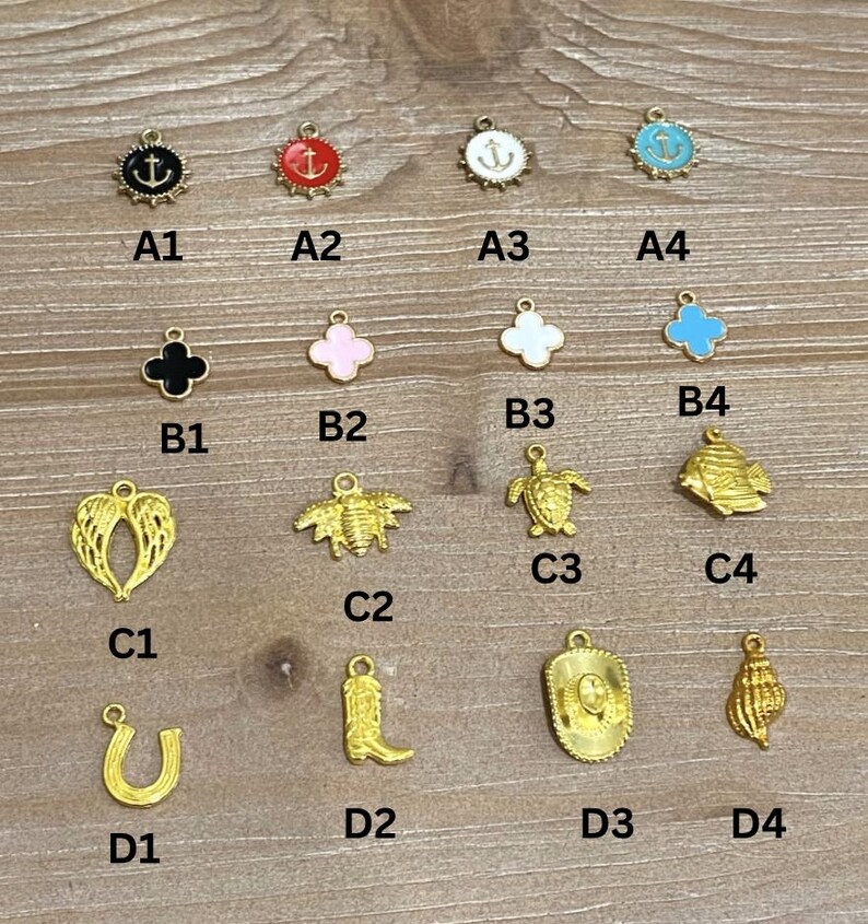 May include: A collection of gold-colored charms with various designs, including an anchor, clover, wings, bee, turtle, fish, horseshoe, cowboy boot, cowboy hat, and seashell. The charms have black, red, white, and blue enamel accents.