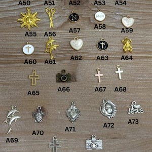 May include: A collection of small metal charms in various shapes, including cows, a sun, scissors, a letter N, hearts, crosses, a teddy bear, a camera, dolphins, a turtle, and a Jesus charm. The charms are displayed on a wooden surface.