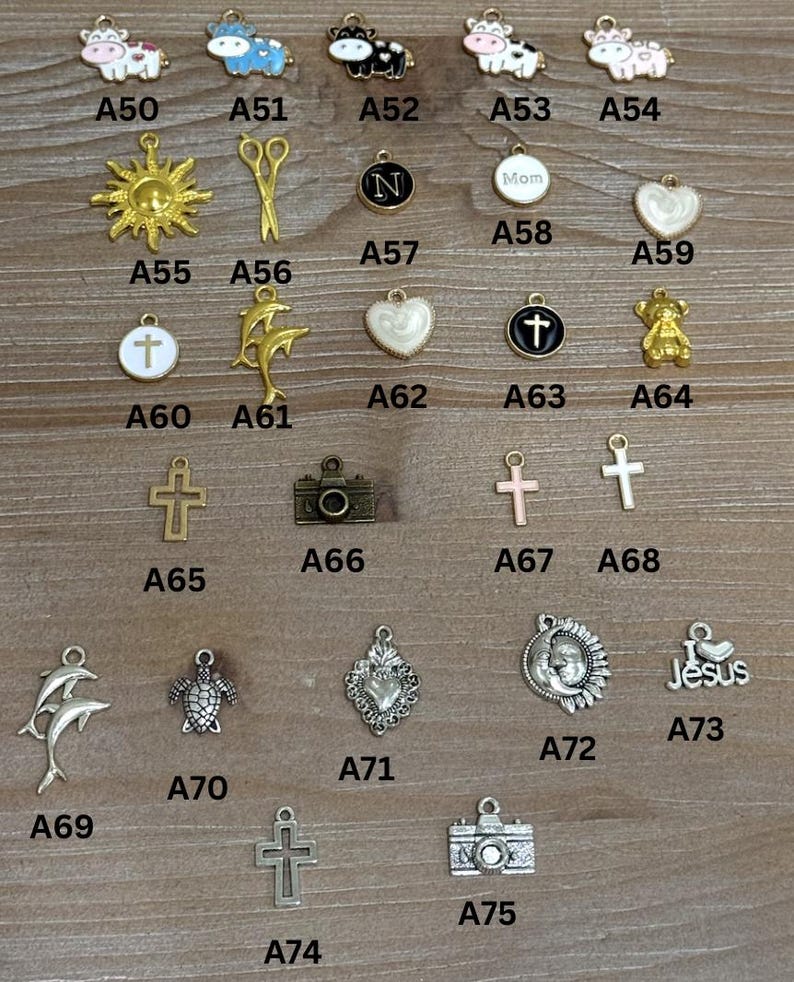May include: A collection of small metal charms in various shapes, including cows, a sun, scissors, a letter N, hearts, crosses, a teddy bear, a camera, dolphins, a turtle, and a Jesus charm. The charms are displayed on a wooden surface.