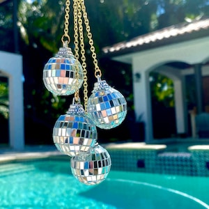 May include: Four small disco ball ornaments hanging from gold chains. The ornaments are silver with mirrored squares, reflecting light. The background includes a swimming pool and a white building.