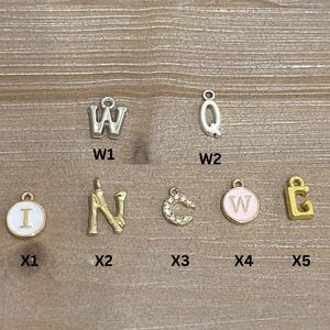 May include: A collection of metal letter charms in various styles. Includes silver and gold-toned letters, some with enamel or rhinestone accents. The letters include W, Q, I, N, C, and E. Ideal for jewelry making or personalization.