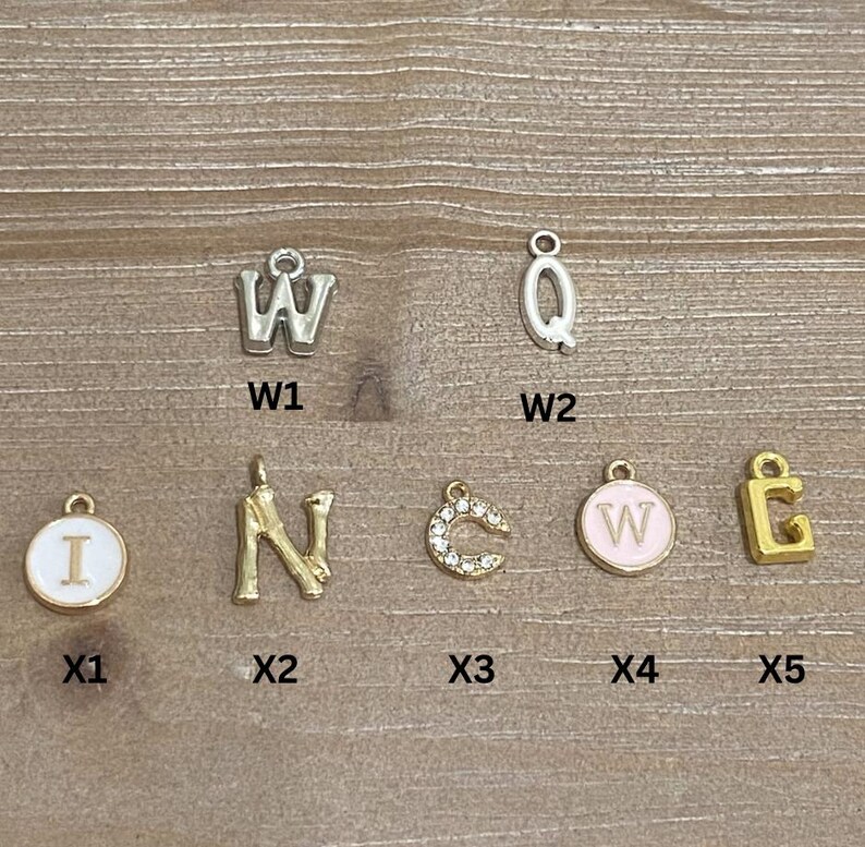 May include: A collection of metal letter charms in various styles. Includes silver and gold-toned letters, some with enamel or rhinestone accents. The letters include W, Q, I, N, C, and E. Ideal for jewelry making or personalization.