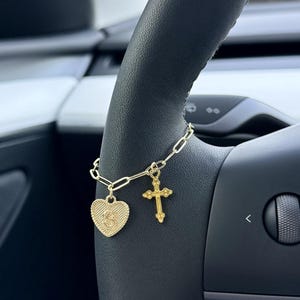 May include: A gold charm bracelet featuring a heart pendant with the letter "S" and a cross pendant. The bracelet is attached to a black steering wheel. The heart pendant has a textured surface.