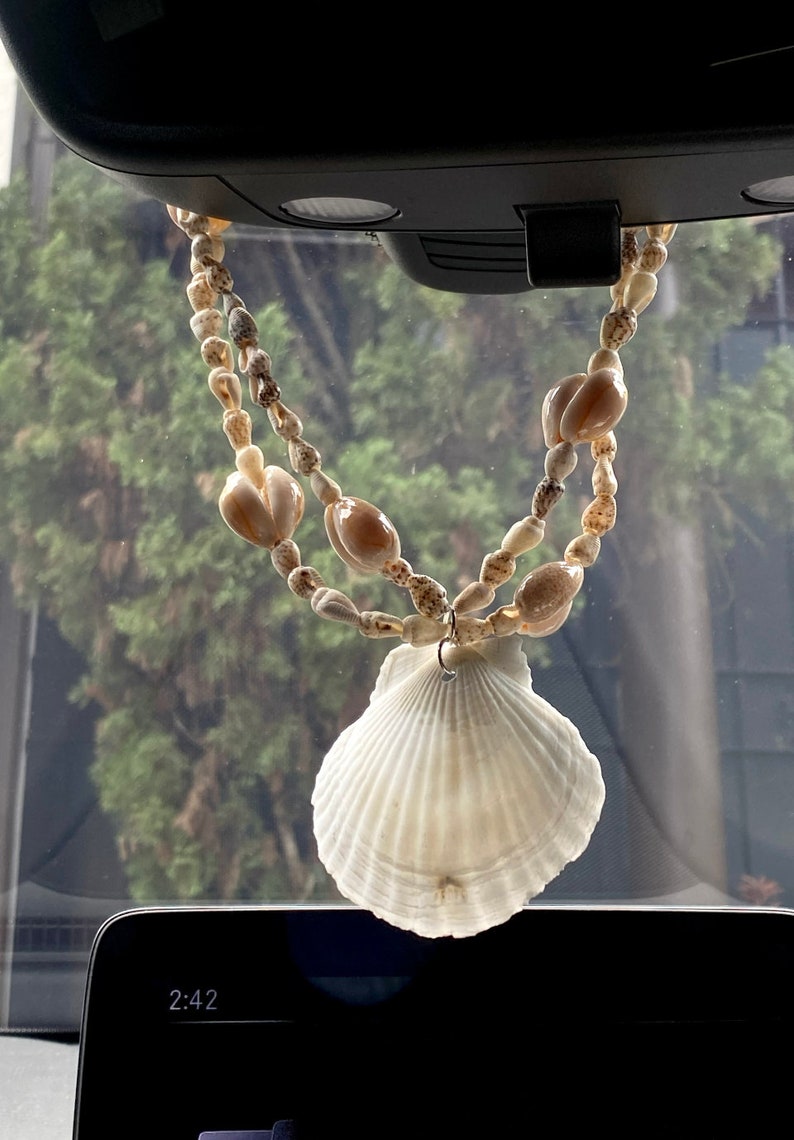 Puede incluir: Una concha blanca colgando del espejo retrovisor de un coche. La concha est&aacute; unida a una cuerda de peque&ntilde;as conchas marrones y blancas.