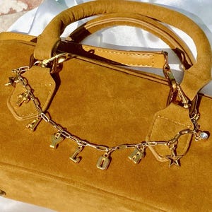 May include: A tan suede handbag with a gold chain and charm detail. The chain spells out "YOLO" in gold letters and includes a star and cross charm. The bag has a rounded handle and a long shoulder strap.