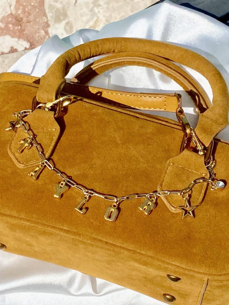 May include: A tan suede handbag with a gold chain and charm detail. The chain spells out "YOLO" in gold letters and includes a star and cross charm. The bag has a rounded handle and a long shoulder strap.