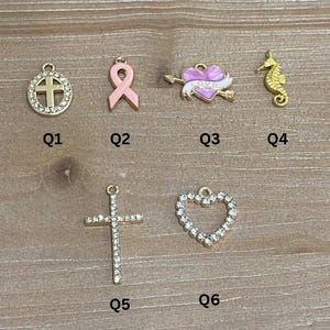 May include: A collection of gold-toned charms, including a cross, a pink ribbon, a heart pierced by an arrow, and a seahorse. Additional charms include a crystal-studded cross and a crystal-lined heart. Each charm has a small loop for attachment.