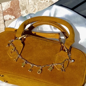May include: Mustard-colored suede handbag with a rounded shape and a top handle. A gold chain with letter charms and star pendants dangles from the handle. The bag has gold hardware and a soft, textured appearance.
