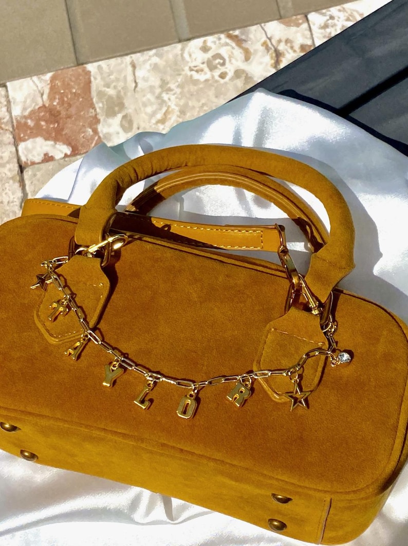 May include: Mustard-colored suede handbag with a rounded shape and a top handle. A gold chain with letter charms and star pendants dangles from the handle. The bag has gold hardware and a soft, textured appearance.