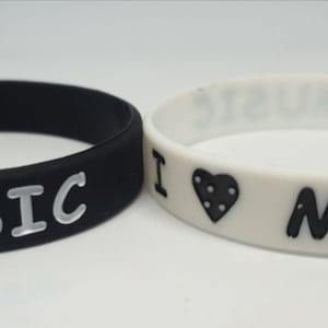 Op de afbeelding: Twee siliconen armbanden, een zwart met witte tekst en een wit met zwarte tekst. Beide armbanden zeggen "Ik hou van muziek" met een hart symbool.