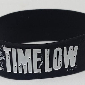 Op de afbeelding: Zwarte rubberen polsband met de tekst "ALL TIME LOW" in witte letters. Een rode schedel en gekruiste botten staan rechts op de band. De band heeft een eenvoudig, cirkelvormig ontwerp.