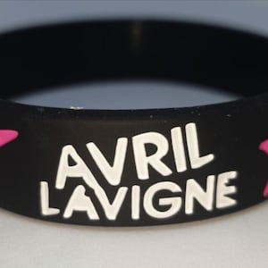 Könnte beinhalten: Schwarzes Gummi-Armband mit dem Namen "AVRIL LAVIGNE" in weißen Blockbuchstaben. Pinkfarbene Sterne flankieren den Text. Ein Accessoire zum Thema Musik.