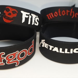 Op de afbeelding: Zwarte rubberen polsbandjes met bandnamen en logo's. Bandjes zijn onder andere "Misfits" met een schedel, "Motorhead", "Lamb of God" en "Metallica". De tekst en logo's zijn in wit en rood.